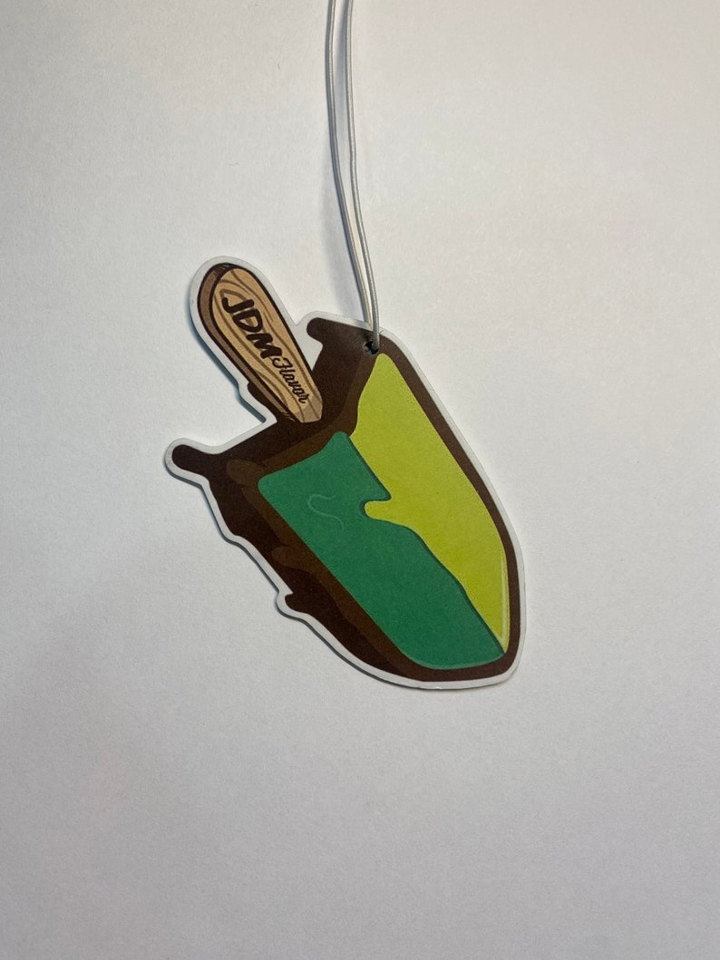 Puede incluir: Un ambientador de coche en forma de paleta verde y amarilla con un palo marr&oacute;n. El palo tiene el texto "JDM Flavors" impreso.