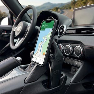 May include: A black 3D-printed smartphone mount installed in a vehicle centre console, featuring the number 124 embossed on its side. The mount holds a smartphone displaying a navigation map with a 12-minute arrival time and route guidance.