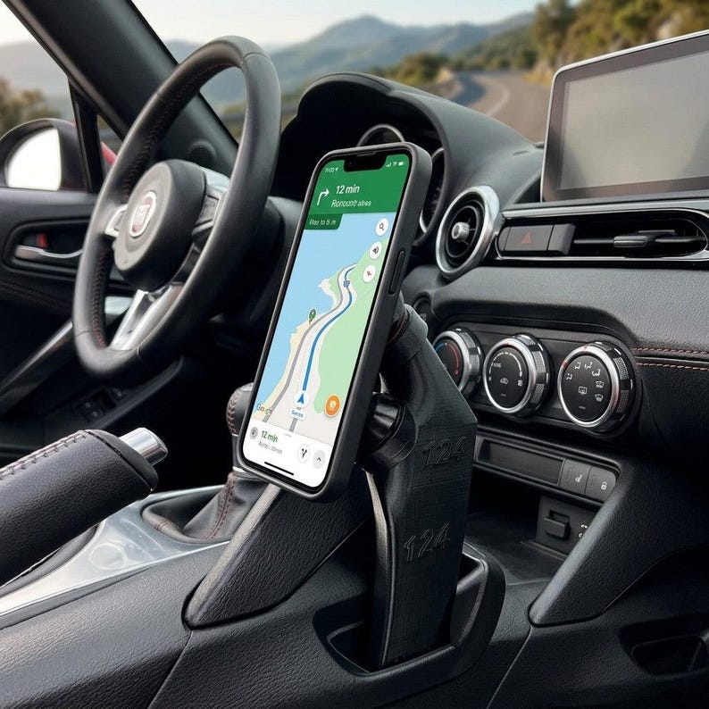 May include: A black 3D-printed smartphone mount installed in a vehicle centre console, featuring the number 124 embossed on its side. The mount holds a smartphone displaying a navigation map with a 12-minute arrival time and route guidance.