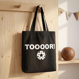 May include: A black tote bag featuring the white text "TOOOOR!" printed above a stylised white football graphic. The bag has two long black shoulder straps and is constructed from a solid fabric material with a simple rectangular design.