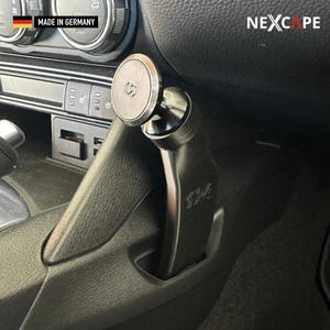 May include: A black magnetic car phone mount installed in the centre console of a vehicle. The mount features a circular magnetic head with a logo, a curved support arm with the text 124, and a base that fits into the console. The text NEXCAPE is visible.