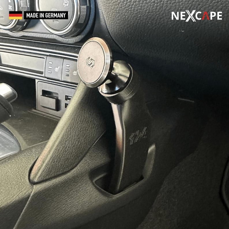 May include: A black magnetic car phone mount installed in the centre console of a vehicle. The mount features a circular magnetic head with a logo, a curved support arm with the text 124, and a base that fits into the console. The text NEXCAPE is visible.