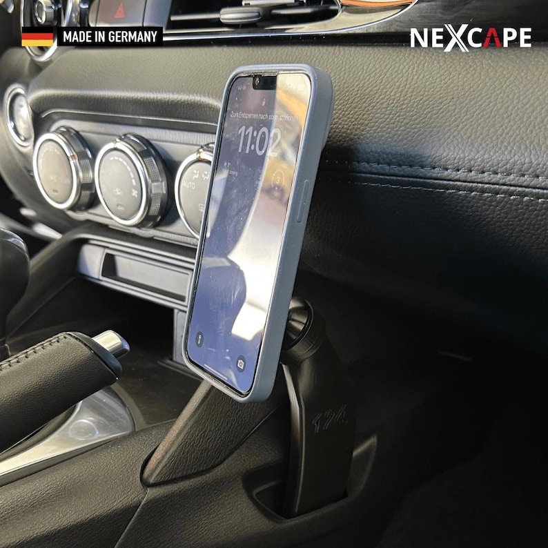 May include: A black car phone mount with a ball joint mechanism holding a smartphone in a light grey case. The mount is secured into the centre console of a vehicle. The text NEXCAPE is visible in the top right corner, and MADE IN GERMANY is present.