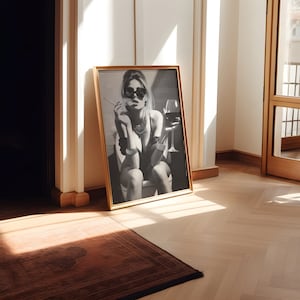 K&ouml;nnte beinhalten: Ein Schwarzwei&szlig;foto einer Frau mit Sonnenbrille und Halskette, die ein Glas Wein und eine Zigarette h&auml;lt. Das Foto ist in einem goldenen Rahmen gerahmt und h&auml;ngt an einer wei&szlig;en Wand.