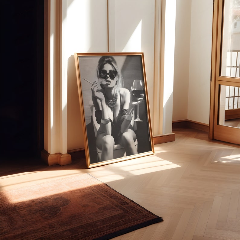 K&ouml;nnte beinhalten: Ein Schwarzwei&szlig;foto einer Frau mit Sonnenbrille und Halskette, die ein Glas Wein und eine Zigarette h&auml;lt. Das Foto ist in einem goldenen Rahmen gerahmt und h&auml;ngt an einer wei&szlig;en Wand.