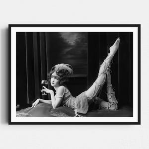 May include: Black and white photograph of a woman in a vintage costume, holding a wine glass. She is lying down with one leg raised, adorned with pearl embellishments. The image evokes a classic, theatrical aesthetic.