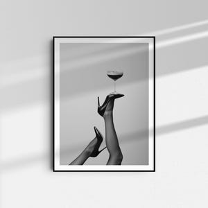 May include: Black and white framed print featuring a pair of legs in black high heels holding a stemmed glass with a dark liquid. The image is a minimalist, artistic representation of fashion and elegance.