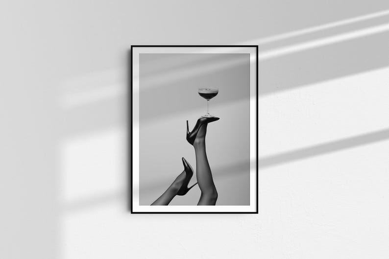 May include: Black and white framed print featuring a pair of legs in black high heels holding a stemmed glass with a dark liquid. The image is a minimalist, artistic representation of fashion and elegance.