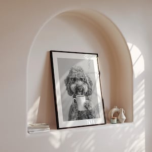 May include: Black and white photo of a dog wearing glasses and holding a coffee mug. The dog is a poodle with a fluffy coat. The photo is framed in a black frame and is hanging on a wall.