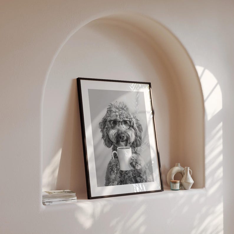 May include: Black and white photo of a dog wearing glasses and holding a coffee mug. The dog is a poodle with a fluffy coat. The photo is framed in a black frame and is hanging on a wall.