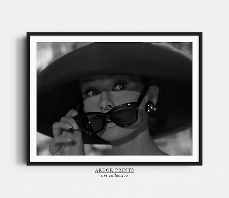 Pu&ograve; includere: Fotografia in bianco e nero di una donna con un cappello grande e occhiali da sole. Sta guardando da un lato e tiene i suoi occhiali da sole con la mano destra.