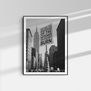 Może przedstawiać: Czarno-biała oprawiona grafika przedstawiająca scenę protestu na ulicy miasta. Ręka trzyma znak z napisem "WE ARE THE DAUGHTERS OF THE WITCHES YOU COULDN'T BURN", z Empire State Building w tle. Obraz jest w czarnej ramie.