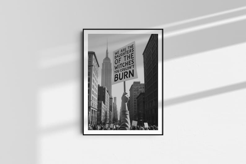 Może przedstawiać: Czarno-biała oprawiona grafika przedstawiająca scenę protestu na ulicy miasta. Ręka trzyma znak z napisem "WE ARE THE DAUGHTERS OF THE WITCHES YOU COULDN'T BURN", z Empire State Building w tle. Obraz jest w czarnej ramie.