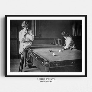 Pode incluir: Duas mulheres jogando bilhar em uma sala com pouca luz. Uma mulher está em pé e dando uma tacada, enquanto a outra mulher está sentada e observando. A mesa de bilhar é de feltro verde com bolas brancas e uma bola preta. A sala tem um piso de madeira e uma mesa de bilhar de madeira escura.