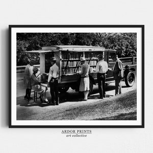 May include: Black and white photograph of a bookmobile, a converted truck with shelves of books, parked on a grassy area. People are browsing and checking out books from the mobile library.