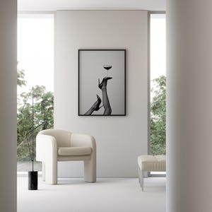 May include: A modern interior scene featuring a framed black and white print of legs in high heels balancing a wine glass. A cream-colored armchair and ottoman sit on a white floor. A black sculpture and large windows complete the minimalist design.
