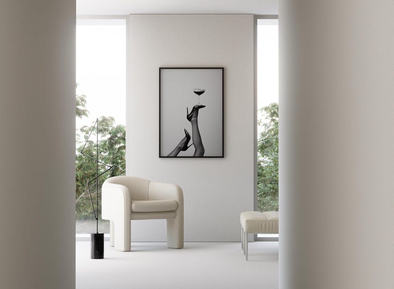 May include: A modern interior scene featuring a framed black and white print of legs in high heels balancing a wine glass. A cream-colored armchair and ottoman sit on a white floor. A black sculpture and large windows complete the minimalist design.