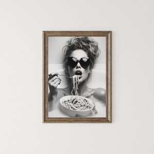 Op de afbeelding: Zwart-witfoto van een vrouw met een zonnebril die spaghetti uit een kom eet in een bad.
