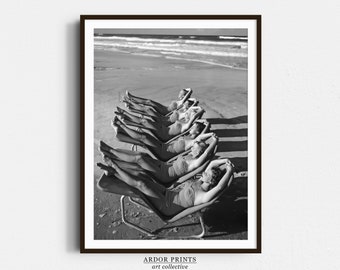 May include: Black and white photograph of a group of women in bathing suits relaxing on deckchairs on a sandy beach. The women are lying on their backs with their arms behind their heads. The sea is in the background.