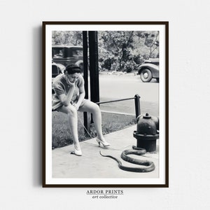 May include: A black and white photograph of a woman sitting on a curb with a snake coiled around a fire hydrant. The woman is wearing a dress and has her chin resting on her hand. The snake is coiled around the fire hydrant, which is labeled "City".