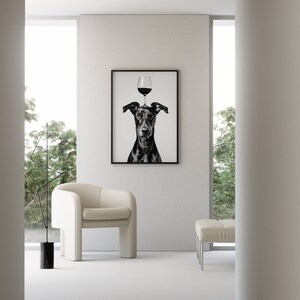 May include: Black and white photograph of a dog with a glass of red wine balanced on its head. The photo is framed and hanging on a white wall in a modern living room.
