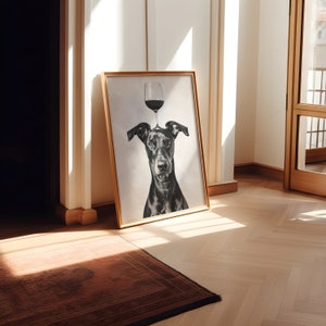 May include: A black and white photo of a dog with a wine glass balanced on its head. The photo is framed in a gold frame and is hanging on a white wall.