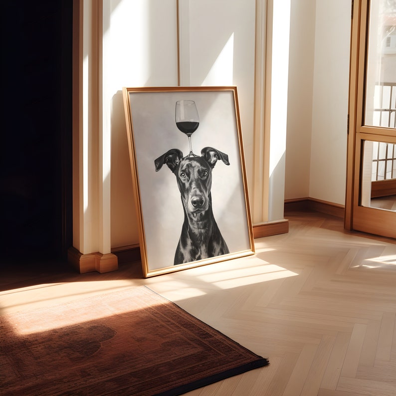 May include: A black and white photo of a dog with a wine glass balanced on its head. The photo is framed in a gold frame and is hanging on a white wall.