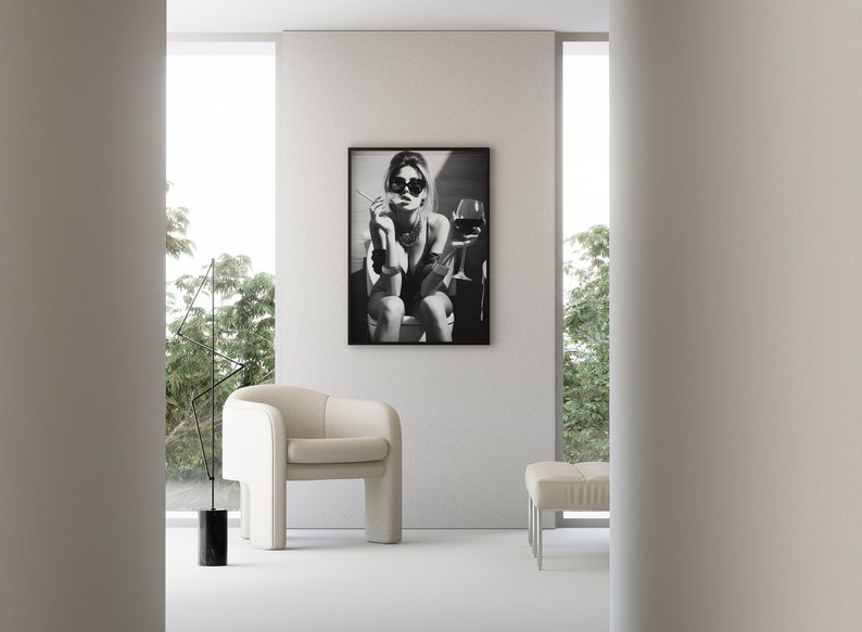 K&ouml;nnte beinhalten: Schwarzwei&szlig;foto einer Frau mit Sonnenbrille, die auf einem Stuhl sitzt und ein Glas Wein und eine Zigarette h&auml;lt. Das Foto ist gerahmt und h&auml;ngt an einer wei&szlig;en Wand.
