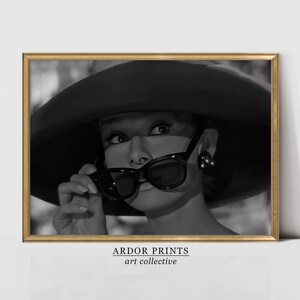 Pu&ograve; includere: Fotografia in bianco e nero di una donna con un grande cappello nero e occhiali da sole. Sta guardando da un lato e ha un'espressione seria sul viso. L'immagine &egrave; incorniciata in una cornice dorata.