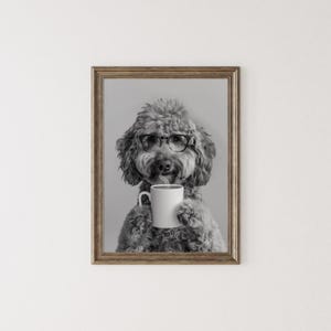 May include: A black and white photo of a dog wearing glasses and holding a white coffee mug. The dog is a poodle with a fluffy coat.
