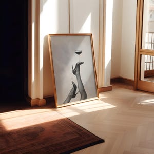 May include: A framed black and white print featuring a glass of red wine balanced on a high-heeled shoe. The artwork is in a gold frame and leans against a wall in a room with hardwood floors and a brown rug.