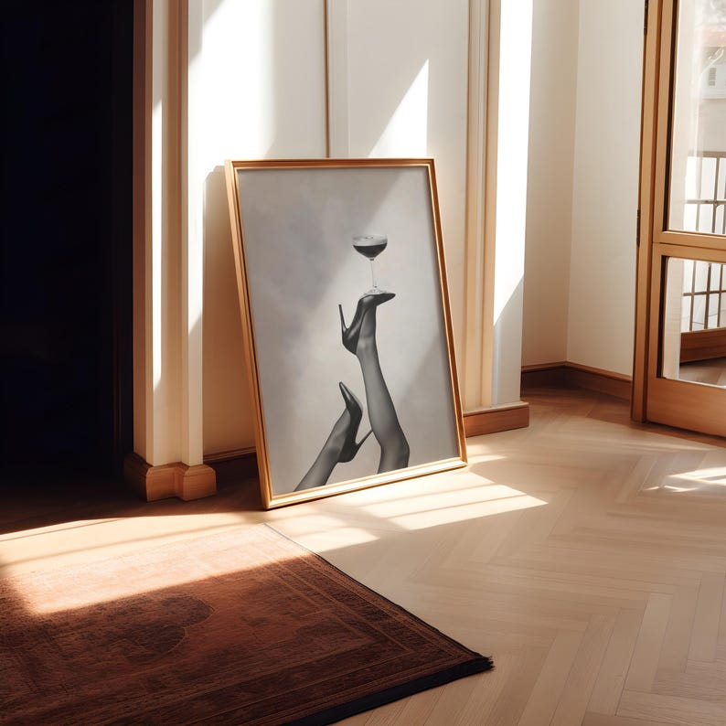 May include: A framed black and white print featuring a glass of red wine balanced on a high-heeled shoe. The artwork is in a gold frame and leans against a wall in a room with hardwood floors and a brown rug.