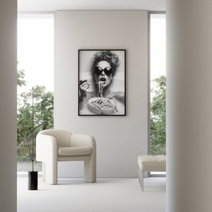 Op de afbeelding: Zwart-witfoto van een vrouw met een zonnebril die spaghetti eet. De foto is ingelijst en hangt aan een witte muur.