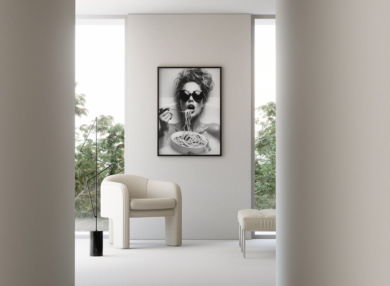 Op de afbeelding: Zwart-witfoto van een vrouw met een zonnebril die spaghetti eet. De foto is ingelijst en hangt aan een witte muur.