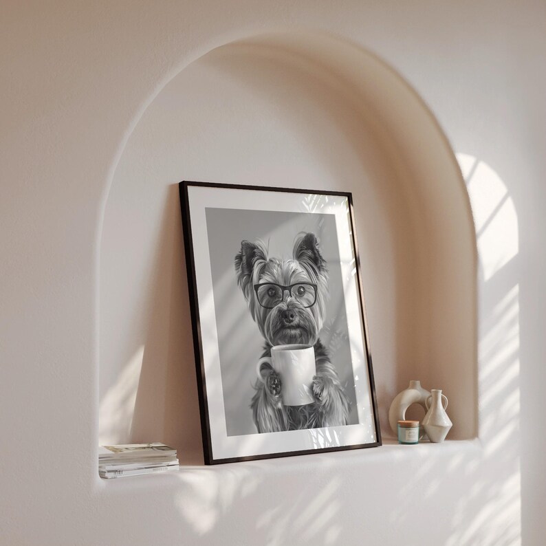 May include: Black and white photograph of a Yorkshire Terrier wearing glasses and holding a white mug. The dog is looking directly at the camera. The photo is framed in a black frame and is hanging on a white wall.