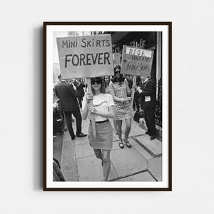 May include: Black and white framed photograph of a protest. A woman in sunglasses holds a sign that reads "Mini Skirts Forever." Other people are in the background, some holding signs. The photograph is taken outdoors on a city street.