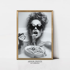 Op de afbeelding: Zwart-witfoto van een vrouw met een zonnebril die spaghetti eet in een badkuip. De foto is ingelijst in een gouden lijst met de tekst "ARDOR PRINTS art collective" onderaan.