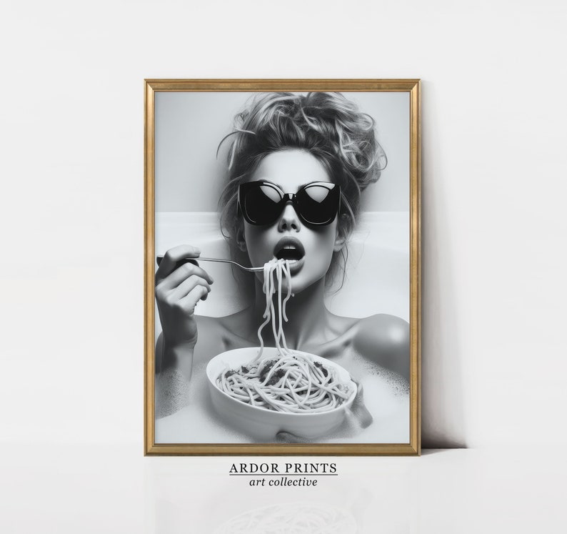 Op de afbeelding: Zwart-witfoto van een vrouw met een zonnebril die spaghetti eet in een badkuip. De foto is ingelijst in een gouden lijst met de tekst "ARDOR PRINTS art collective" onderaan.