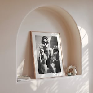 K&ouml;nnte beinhalten: Schwarzwei&szlig;foto einer Frau mit Sonnenbrille und schwarzem Oberteil, die auf einer Toilette sitzt und ein Glas Wein h&auml;lt. Das Foto ist in einem Holzrahmen gerahmt.