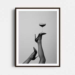 May include: Black and white framed print featuring a cocktail glass balanced on a high-heeled shoe. The image showcases a pair of legs in sheer stockings, with one leg extended upwards, holding the glass. The overall aesthetic is elegant and stylish.