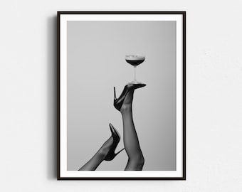 May include: Black and white framed print featuring a cocktail glass balanced on a high-heeled shoe. The image showcases a pair of legs in sheer stockings, with one leg extended upwards, holding the glass. The overall aesthetic is elegant and stylish.