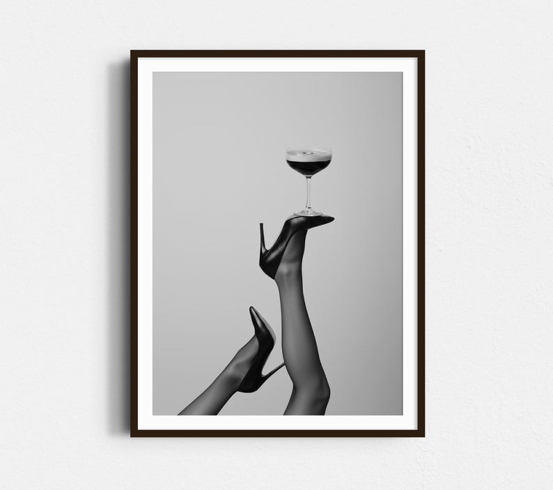 May include: Black and white framed print featuring a cocktail glass balanced on a high-heeled shoe. The image showcases a pair of legs in sheer stockings, with one leg extended upwards, holding the glass. The overall aesthetic is elegant and stylish.