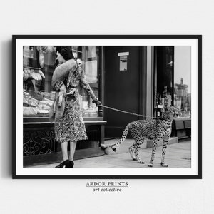 May include: A black and white photograph of a woman in a patterned dress walking a cheetah on a lead in front of a shop. The woman is wearing a fur stole and is looking to the side. The cheetah is walking calmly beside her.