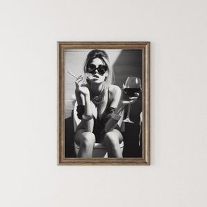 K&ouml;nnte beinhalten: Schwarzwei&szlig;foto einer Frau mit Sonnenbrille und schwarzem Oberteil, die auf einer Toilette sitzt, ein Glas Rotwein h&auml;lt und eine Zigarette raucht.