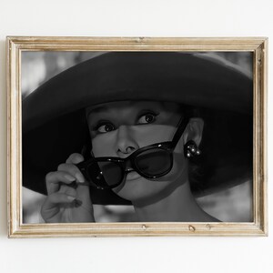 Pu&ograve; includere: Foto in bianco e nero di una donna con un cappello grande e occhiali da sole. Sta guardando da un lato e tiene gli occhiali da sole con la mano destra.