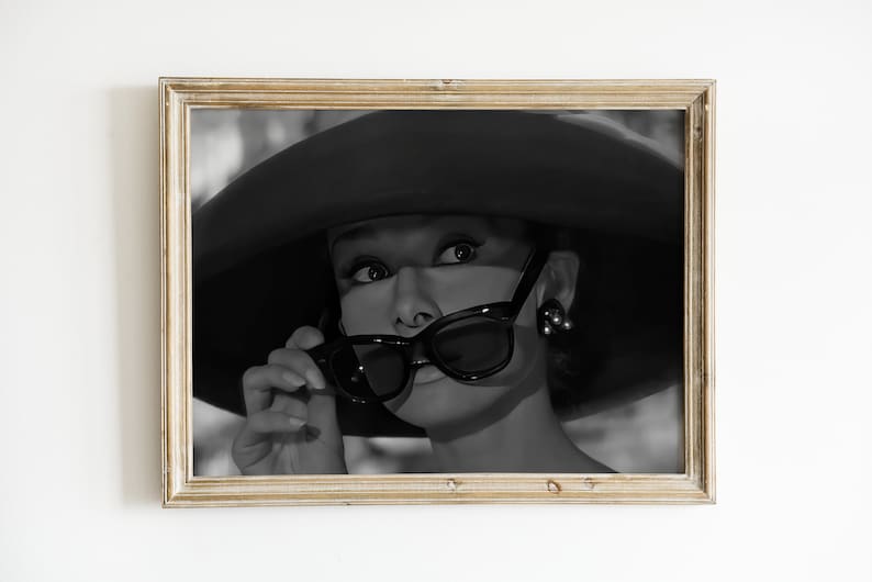Pu&ograve; includere: Foto in bianco e nero di una donna con un cappello grande e occhiali da sole. Sta guardando da un lato e tiene gli occhiali da sole con la mano destra.