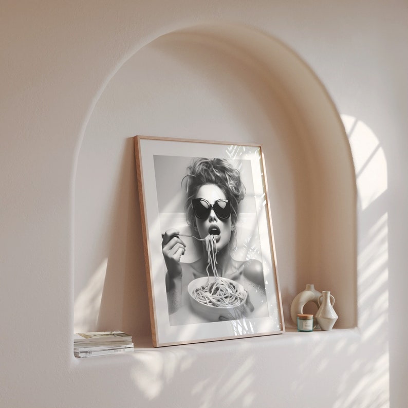 Op de afbeelding: Zwart-witfoto van een vrouw met een zonnebril die spaghetti eet. De foto is ingelijst in een houten lijst.