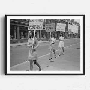 May include: A black and white photograph showing three people walking down a street, each carrying a sign. The signs read "MINI SKIRTS FOREVER!" and "WE SUPPORT THE MINI SKIRT". The people are wearing mini skirts and dresses.