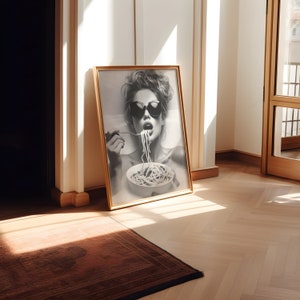 Op de afbeelding: Een zwart-witfoto van een vrouw met een zonnebril die spaghetti eet. De foto is ingelijst in een gouden lijst en hangt aan een muur.