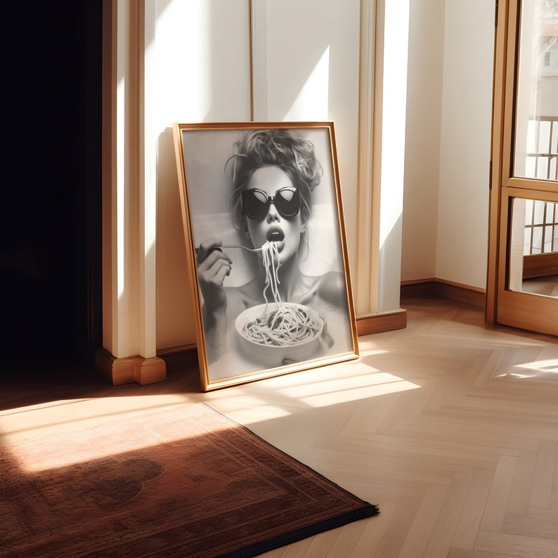 Op de afbeelding: Een zwart-witfoto van een vrouw met een zonnebril die spaghetti eet. De foto is ingelijst in een gouden lijst en hangt aan een muur.